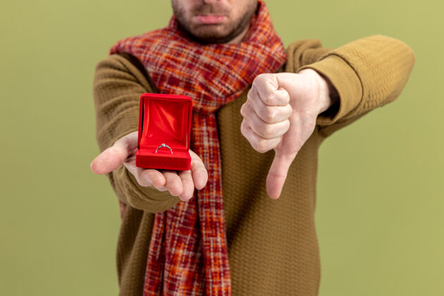 拇指穿着休闲服的年轻人脖子上围着围巾 红色盒子上戴着订婚戒指 拇指朝下 情人节的概念站在绿色背景上下围巾戒指