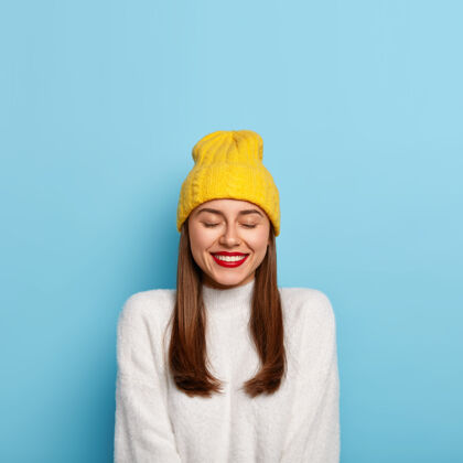 寒冷可爱的黑发女人 妆最少 红唇膏 笑得愉快 戴着黄色帽子和白色跳线 隔着蓝色的墙人们欢呼肖像