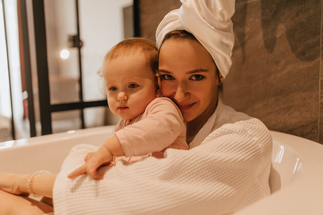 淋浴一个穿着白色长袍 头上戴着毛巾的女士的特写肖像 她和女儿坐在白色的深浴盆里妈妈女性浴袍