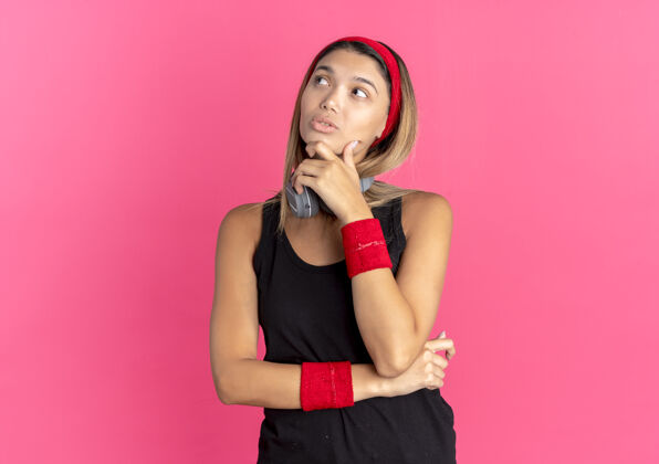 黑色身穿黑色运动服 头戴红色耳机 手放在下巴上 面带粉色困惑的年轻健身女孩立场手头带