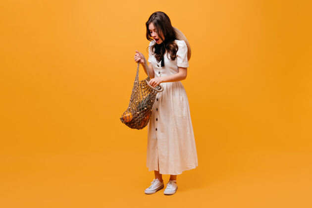 线包一位身穿白色连衣裙的女士打开装满水果的购物袋 在橙色背景上摆出姿势卷发姿势瘦身