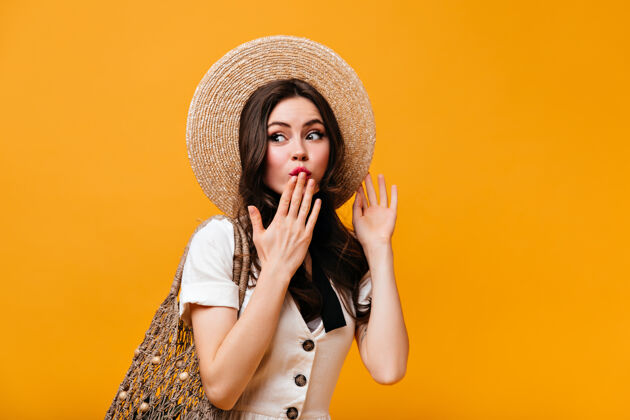 女性戴草帽 穿白色t恤 手捂嘴 拿着橙色背景购物袋的调情女人的画像卷发黑发瘦身