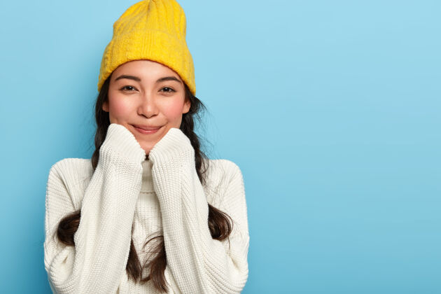 肖像快乐可爱的亚洲女人的画像 手放在下巴下 穿着超大的白色毛衣和黄色帽子 享受舒适和平静的家庭气氛温暖微笑民族
