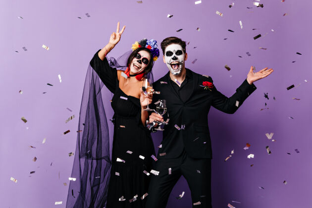 骷髅身着万圣节服装的活跃男女在五彩纸屑中的紫色背景上翩翩起舞香槟墨西哥节日