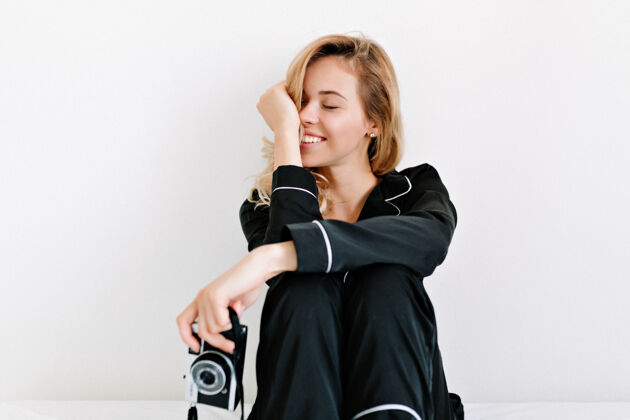 女士室内摄影棚拍摄的可爱快乐的女人闭上眼睛 微笑 而坐在床上的白色墙壁与复古相机相机房子成人