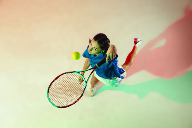 活动身穿蓝色球衣的年轻女子正在打网球她用球拍击球室内射门 光线混合年轻 灵活 力量和活力顶视图专业私人教练休闲