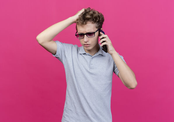 马球一个戴着黑眼镜 穿着灰色马球衫的失望的年轻人站在粉色的墙上 一边用手机聊天 一边摸着头 心里很困惑说话头穿