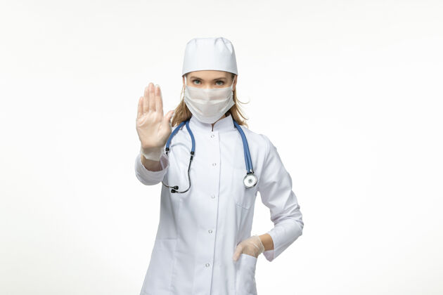 佩戴正面图女医生身穿医疗服戴口罩因冠状病毒在白墙上显示停止标志疾病冠状病毒-大流行病毒疾病医生女性专业