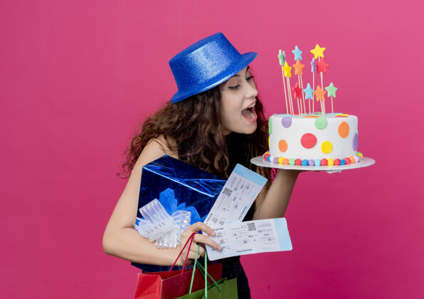 蛋糕一位年轻漂亮的女士 卷发 戴着节日礼帽 手里拿着生日蛋糕礼盒和机票 站在粉色的墙上 快乐而兴奋的生日派对概念机票女人帽子
