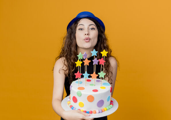 抱着一个年轻漂亮的女人 卷发 戴着节日礼帽 手里拿着生日蛋糕 快乐而积极地站在橘色的墙上亲吻年轻吹蛋糕