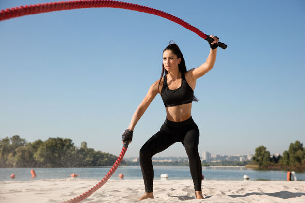 私人教练年轻健康的女人在沙滩上用绳子锻炼身体健康蹲肌肉