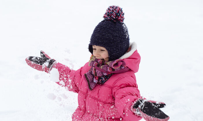 冬天一个穿着粉色夹克 戴着帽子的可爱小女孩正在雪地里玩耍冬季儿童娱乐理念玩孩子游戏