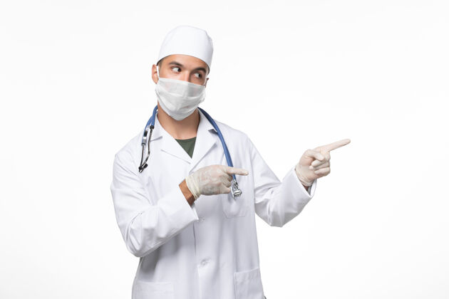 疾病正面图：男医生穿着医疗服 戴着防毒面具 白色办公桌上带着蓝色听诊器的科维德-疾病大流行病毒视图办公桌防护