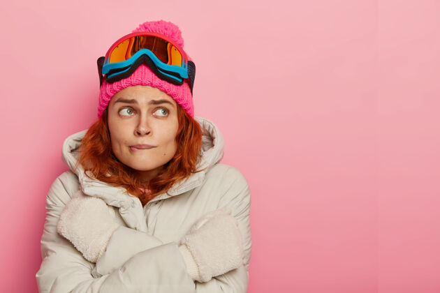 外套滑雪者感到寒冷 颤抖和咬嘴唇 穿着温暖的冬季外套 试图温暖 站在粉红色的工作室墙 复制空间女人冬天滑雪