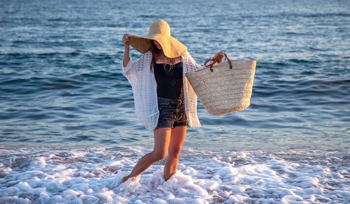 长廊一个戴着大帽子和柳条袋的女孩沿着海边散步暑假概念海岸散步赤脚