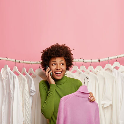 粉色快乐的非洲裔美国女人在展厅里挑选新衣服 拿起衣架上的紫色高领毛衣 站在衣架上的白色衣服上面时尚手机高领毛衣
