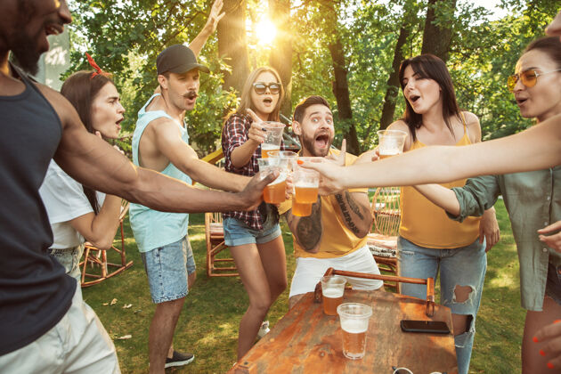休闲一群快乐的朋友在阳光明媚的日子里喝着啤酒 一起烧烤一起在户外的林间空地或后院休息家庭饮料啤酒