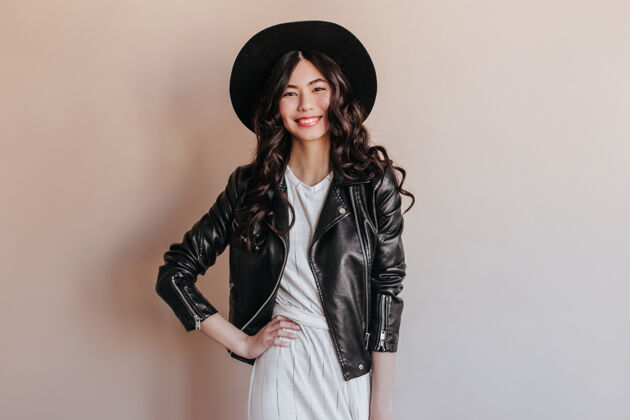肖像穿着皮夹克的亚洲女人站着 手放在臀部摄影棚拍摄的微笑的中国女人戴着帽子 背景是米色的享受中国人日本人