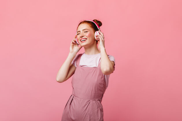肖像一个穿着浅色工作服的开朗的女人在粉色背景下跳舞 戴着耳机听音乐粉色背景衬衫优雅