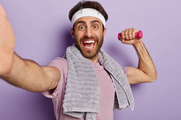 强壮精力充沛的欧洲运动员开怀大笑 用哑铃锻炼肱二头肌 自拍肖像 脖子上围着毛巾 戴着头带健身单身微笑