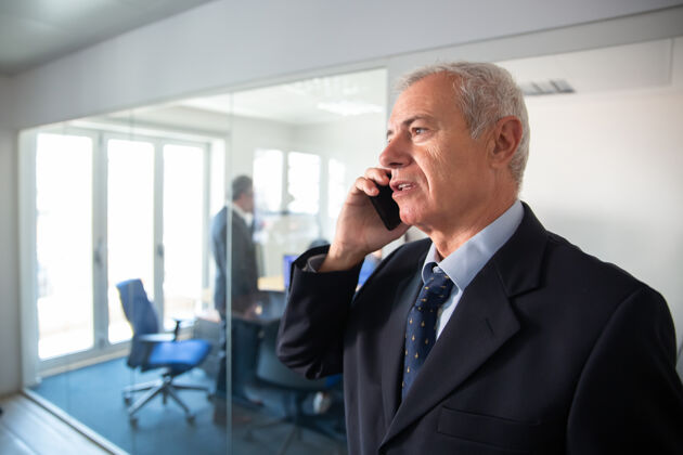 公司专注的成熟商人在办公室玻璃墙上讲手机 站在走廊里沟通理念年龄财务会议