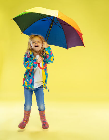 可爱黄色工作室墙上 一个穿着雨衣 手持彩虹色雨伞的时髦女孩的完整肖像美丽肖像多彩
