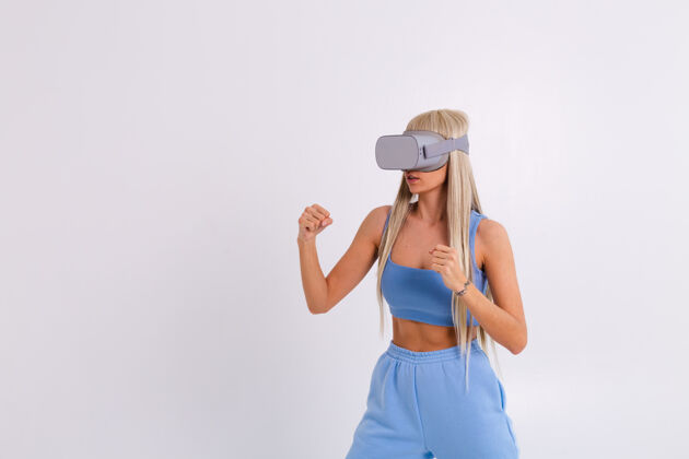 模拟摄影棚照片中 一个年轻漂亮的女子穿着温暖的蓝色时尚西装 戴着虚拟现实眼镜 在白色舞台上打拳击游戏模仿情感