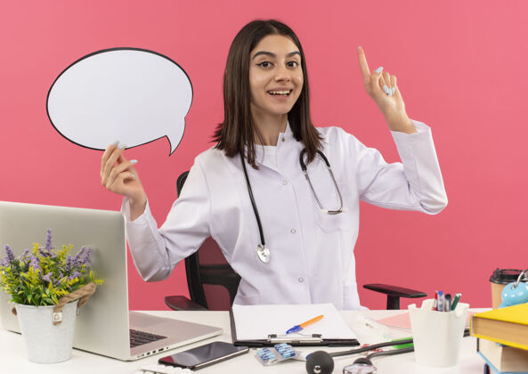 坐着年轻的女医生穿着白大褂 脖子上戴着听诊器 手里拿着空白的语音气泡 手指朝上 坐在桌边 笔记本电脑放在粉色的墙上笔记本电脑脖子泡泡