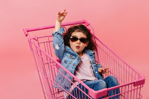 情绪有趣的孩子戴着太阳镜坐在购物车里摄影棚拍摄了一个穿着牛仔夹克的快乐小女孩表情肖像玩