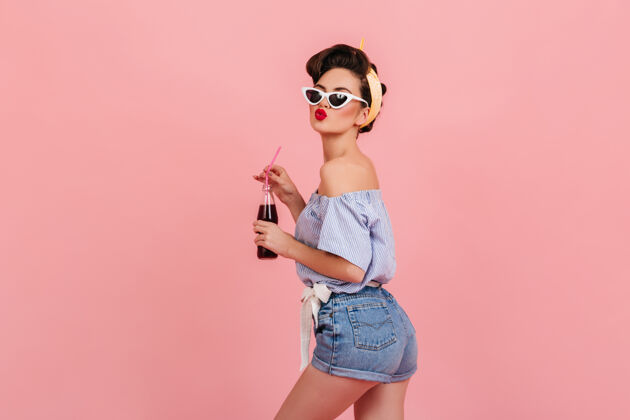 优雅可爱的小妞穿着牛仔短裤表达积极的情绪摄影棚拍摄了一位戴着太阳镜的快乐黑发女士拿着粉红色背景的饮料瓶衬衫瓶子女性