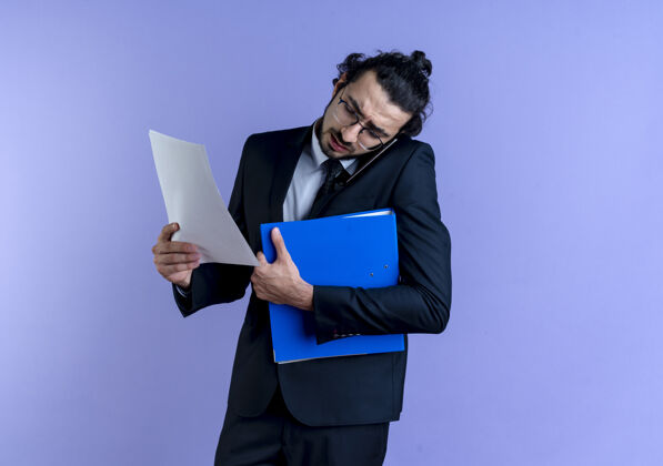 商务一个穿着黑西装戴眼镜的商人站在蓝色的墙上 手里拿着文件夹和文件 一边用手机聊天眼镜举行文件
