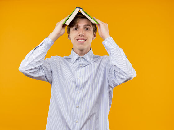 微笑一个身穿白衬衫的年轻人头顶着一本打开的书 站在橘色的墙上开心地笑着开放抱着站着