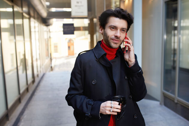 交谈积极的年轻迷人的棕色头发的男性 穿着时髦的衣服 拿着外卖咖啡 一边用智能手机打电话 在城市背景中漫步衣服电话男性