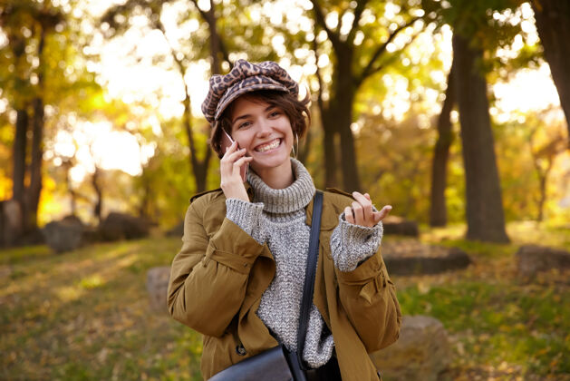 户外在秋日阳光明媚的日子里 一位年轻迷人的快乐短发黑发女子在公园散步 一边愉快地看着前方 一边灿烂地微笑着 一边愉快地打电话秋天白天手机