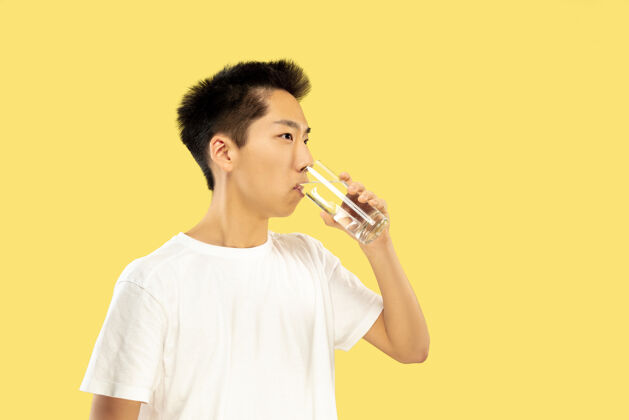 手势黄色摄影棚背景上的韩国年轻人半身像穿白衬衫的男模饮用水人类情感的概念 面部表情正面视图流行色彩男人男性工作