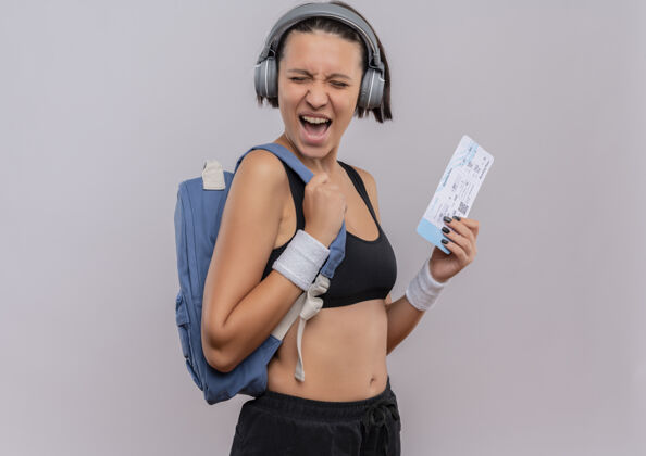 快乐年轻的健身女 头戴耳机 背着背包 手里拿着机票 站在白色的墙上 疯狂地高喊着疯狂喊叫票