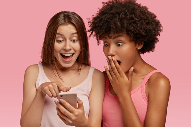 混合两个多民族的年轻女孩通过手机查看网页上的精彩文章 有着惊喜的快乐表情 穿着夏装 使用无线网络 模特对着粉色的墙壁室内指向青少年