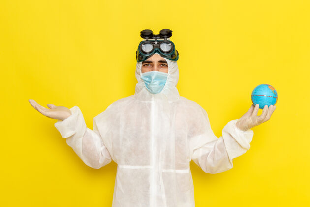 服装正面图：黄色办公桌上 一位身着特殊套装的男科学工作者手里拿着一个小圆球化学保护套套装