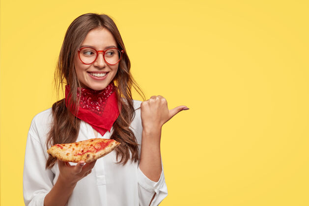 视图一张迷人的微笑女孩的照片向比萨店指明了方向 她吃着美味的比萨配奶酪和西红柿 用拇指指着靠着黄色墙壁的复制空间女人在室内吃零食快餐件烹饪
