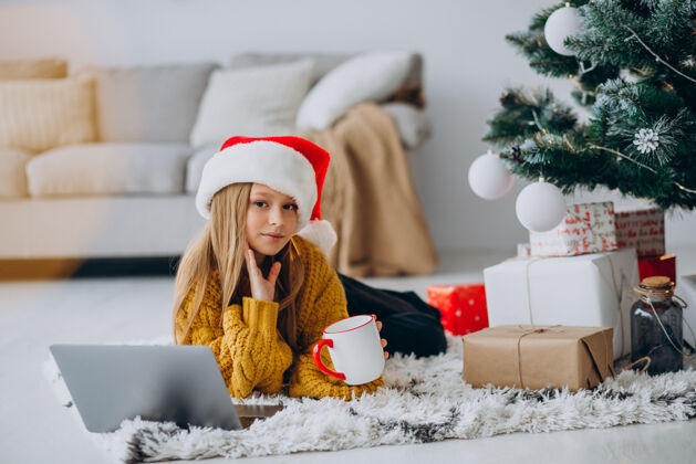 电脑可爱的女孩在圣诞树旁用电脑科技饮酒圣诞树