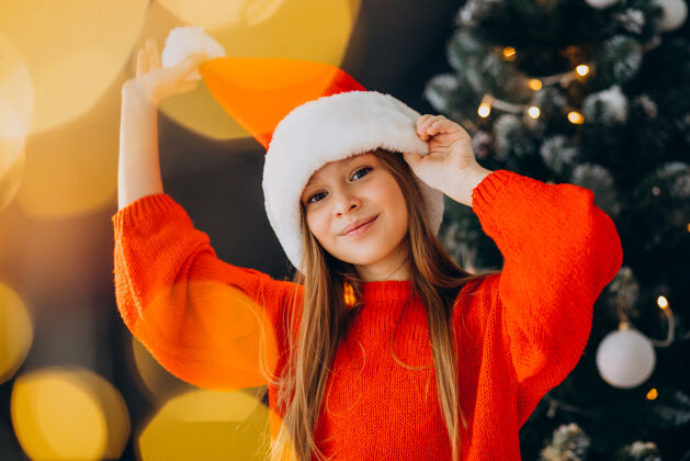 积极可爱的少女戴着圣诞树旁的红色圣诞帽圣诞树时尚红毛衣