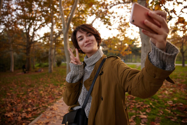 城市积极向上的年轻可爱的黑发短发女子一边用手机拍照 一边用和平的手势举手 在城市花园里的黄树上摆姿势树女士温暖