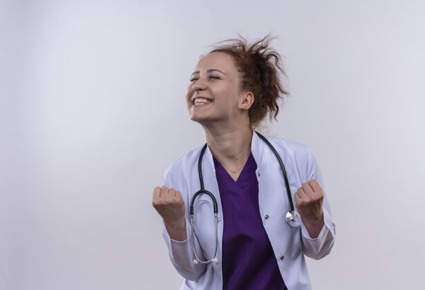 幸福年轻的女医生穿着白大褂 手持听诊器 握紧拳头 欢欣鼓舞地站在白墙上高兴年轻拳头