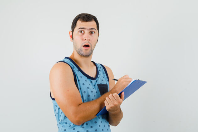 自信一个年轻人穿着蓝色的单件衣服在剪贴板上做笔记 他看起来很惊讶 正视图欢呼时尚聪明