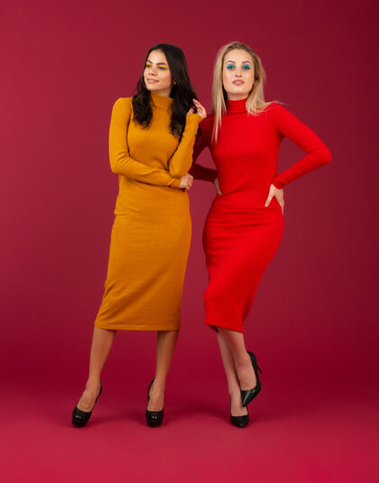 毛衣两位穿着黄色和红色秋冬时尚针织连衣裙的迷人时尚女性在红墙上孤立地摆着姿势微笑魅力女性