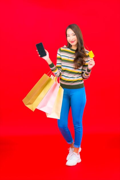手机在红墙上画一幅美丽的亚洲年轻女子的画像 上面有五颜六色的购物袋和信用卡销售借记购物狂