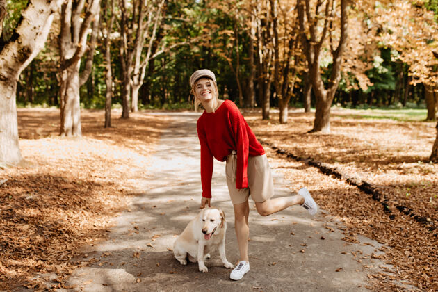 朋友穿着时尚的红色套头衫和米色短裤的快乐金发女郎和她的狗在美丽的秋天公园里玩得很开心散步户外成人