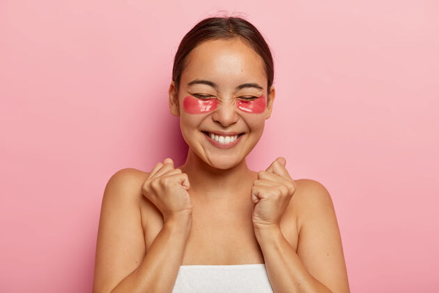 女性治疗眼底 皮肤护理快乐满意的亚洲女人有眼底的化妆品贴片 以减少浮肿 握紧拳头从喜悦和快乐 有美容程序在家水疗黑发姿势
