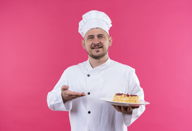 盘子面带微笑的年轻帅哥厨师身着厨师制服拿着一盘蛋糕指着它 手孤立在粉红色的空间里帅气年轻蛋糕