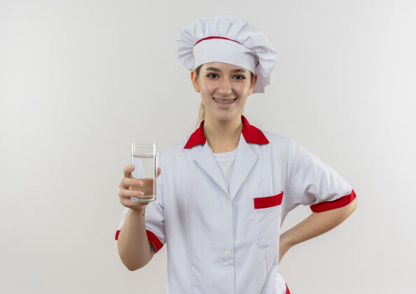 漂亮面带微笑的年轻漂亮厨师 身着厨师制服 戴着牙套 端着一杯水 把手放在腰部 与白色空间隔离开来微笑厨师水